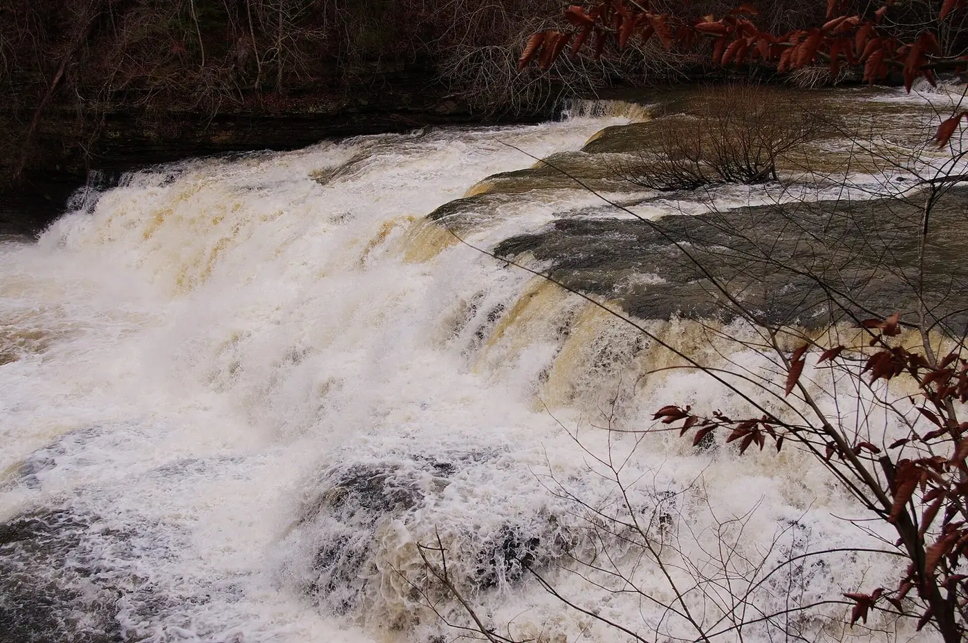 Brown floodwater rushing over a wide, low rock ledge on the Falling Water River
